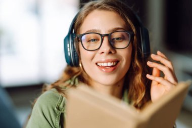 Kablosuz kulaklık takan ve güneş ışığında kitap tutan genç bir kadının yakın çekimi. Gözlüklü, gülümseyen bir kadın kitap okuyor ve rahat bir odada müzik dinlemekten hoşlanıyor. Konsept: eğitim, teknoloji.