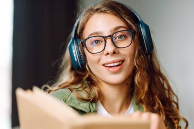 Kablosuz kulaklık takan ve güneş ışığında kitap tutan genç bir kadının yakın çekimi. Gözlüklü, gülümseyen bir kadın kitap okuyor ve rahat bir odada müzik dinlemekten hoşlanıyor. Konsept: eğitim, teknoloji.