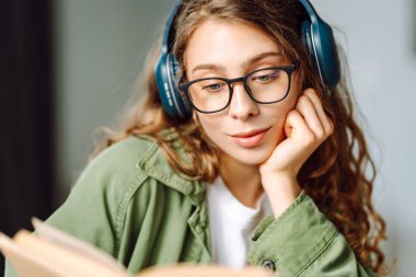 Kablosuz kulaklık takan ve güneş ışığında kitap tutan genç bir kadının yakın çekimi. Gözlüklü, gülümseyen bir kadın kitap okuyor ve rahat bir odada müzik dinlemekten hoşlanıyor. Konsept: eğitim, teknoloji.