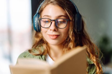 Kablosuz kulaklık takan ve güneş ışığında kitap tutan genç bir kadının yakın çekimi. Gözlüklü, gülümseyen bir kadın kitap okuyor ve rahat bir odada müzik dinlemekten hoşlanıyor. Konsept: eğitim, teknoloji.