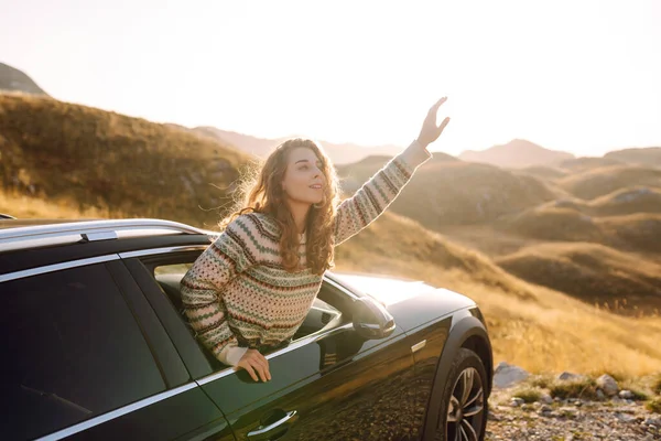 Mutlu bir gezgin gün batımında arabanın camından sarkar. Genç kadın kendini özgür hissediyor ve araba sürerken dağ manzarasının tadını çıkarıyor. Macera ve ulaşım konsepti. Etkin yaşam biçimi.