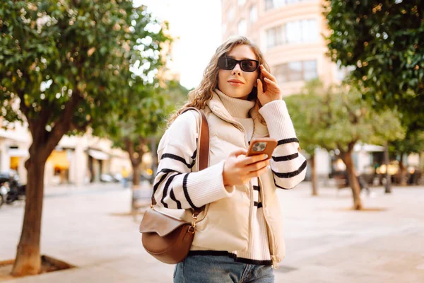 Güneş gözlüklü genç bir kadın telefonuyla şehrin güneşli sokaklarında geziniyor. Bir turist gün batımının tadını çıkarıyor, telefonundan sosyal medyayı kontrol ediyor. Konsept: Turizm, yürüyüş.