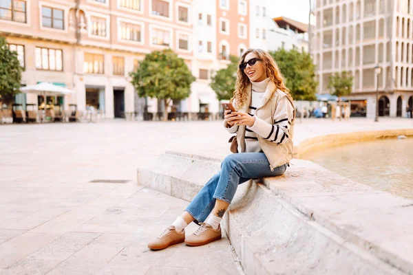 Güneş gözlüklü genç bir kadın telefonuyla şehrin güneşli sokaklarında geziniyor. Bir turist gün batımının tadını çıkarıyor, telefonundan sosyal medyayı kontrol ediyor. Konsept: Turizm, yürüyüş.