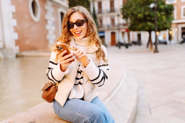 Güneş gözlüklü genç bir kadın telefonuyla şehrin güneşli sokaklarında geziniyor. Bir turist gün batımının tadını çıkarıyor, telefonundan sosyal medyayı kontrol ediyor. Konsept: Turizm, yürüyüş.