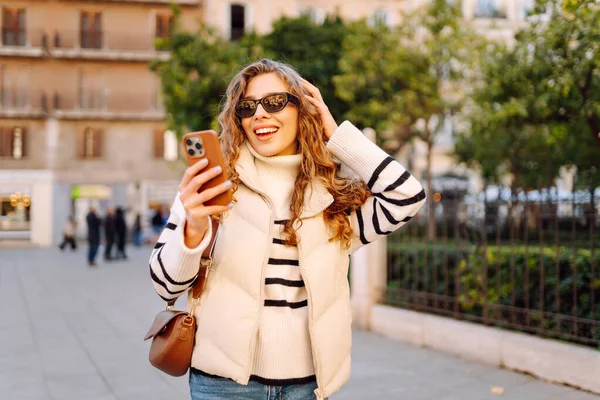 Güneş gözlüklü genç bir kadın telefonuyla şehrin güneşli sokaklarında geziniyor. Bir turist gün batımının tadını çıkarıyor, telefonundan sosyal medyayı kontrol ediyor. Konsept: Turizm, yürüyüş.