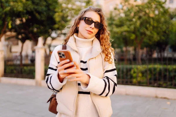 Güneş gözlüklü genç bir kadın telefonuyla şehrin güneşli sokaklarında geziniyor. Bir turist gün batımının tadını çıkarıyor, telefonundan sosyal medyayı kontrol ediyor. Konsept: Turizm, yürüyüş.