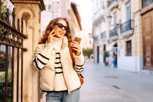 Güneş gözlüklü genç bir kadın telefonuyla şehrin güneşli sokaklarında geziniyor. Bir turist gün batımının tadını çıkarıyor, telefonundan sosyal medyayı kontrol ediyor. Konsept: Turizm, yürüyüş.