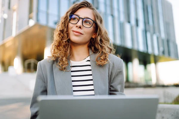 Şık giyinmiş bir kadın basamaklarda dizüstü bilgisayarla modern bir binanın arka planına karşı oturur. Güzel bayan serbest yazar dizüstü bilgisayarda çalışıyor ve güneşli günün tadını çıkarıyor. Uzak iş ve iş için kavram.