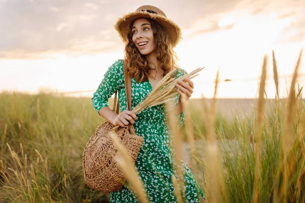 Uzun hasır şapkalı gülümseyen bir kadın sahile yakın poz veriyor. Saman çantalı güzel bir kadın deniz manzarasının ve gün batımının tadını çıkarıyor. Kavram: doğa, güzellik, yaşam tarzı.