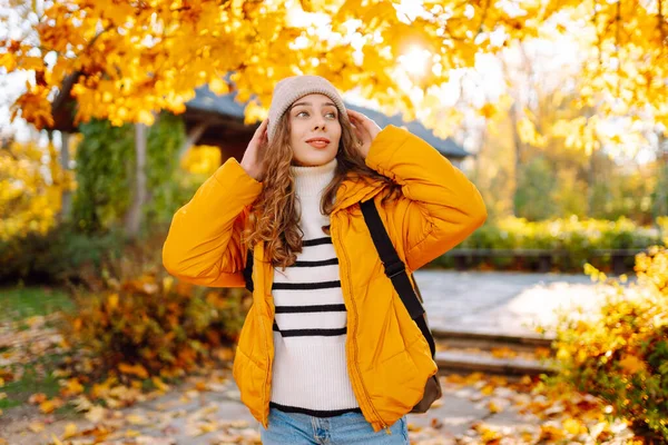Sırt çantası ve parlak ceketli genç bir gezginin portresi sonbahar parkında gün batımında geziniyor. Mutlu kadın ormanda eğleniyor ve güneşli bir günün tadını çıkarıyor. Seyahat ve hafta sonu kavramı.