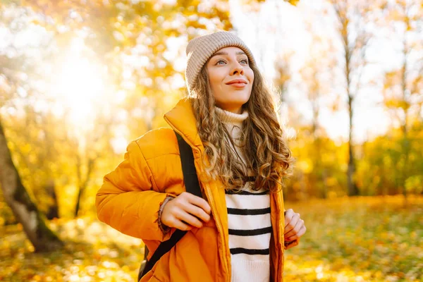 Sırt çantası ve parlak ceketli genç bir gezginin portresi sonbahar parkında gün batımında geziniyor. Mutlu kadın ormanda eğleniyor ve güneşli bir günün tadını çıkarıyor. Seyahat ve hafta sonu kavramı.