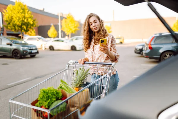 Güzel bir kadın, arabasının yanındaki güneşli otoparkta taze meyve ve sebzelerle duruyor. Elinde alışveriş arabası ve poşet dolusu yiyecek olan bir kadın gün batımının tadını çıkarır. Kavram tutumlu ve tazedir..