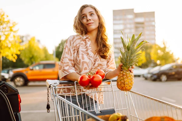 Güzel bir kadın, arabasının yanındaki güneşli otoparkta taze meyve ve sebzelerle duruyor. Elinde alışveriş arabası ve poşet dolusu yiyecek olan bir kadın gün batımının tadını çıkarır. Kavram tutumlu ve tazedir..