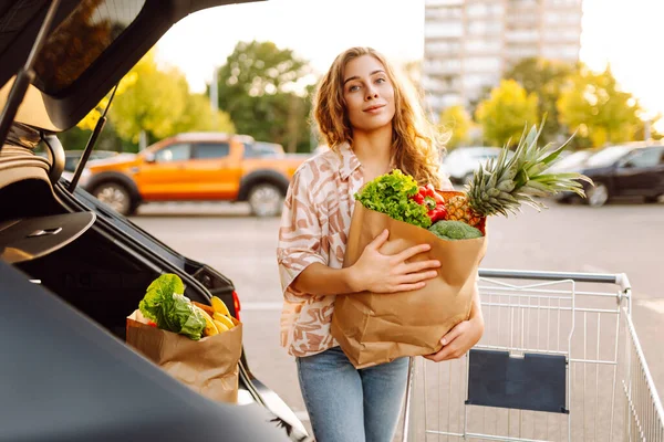 Güzel bir kadın, arabasının yanındaki güneşli otoparkta taze meyve ve sebzelerle duruyor. Elinde alışveriş arabası ve poşet dolusu yiyecek olan bir kadın gün batımının tadını çıkarır. Kavram tutumlu ve tazedir..