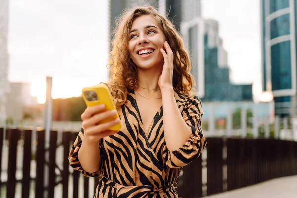 Elinde telefon olan genç bir kadın şehir manzarasının arka planında bir sokak arasında geziniyor. Güzel bir kadın telefonuyla güneşli bir günün tadını çıkarır. Teknoloji konsepti. Yaşam biçimi.