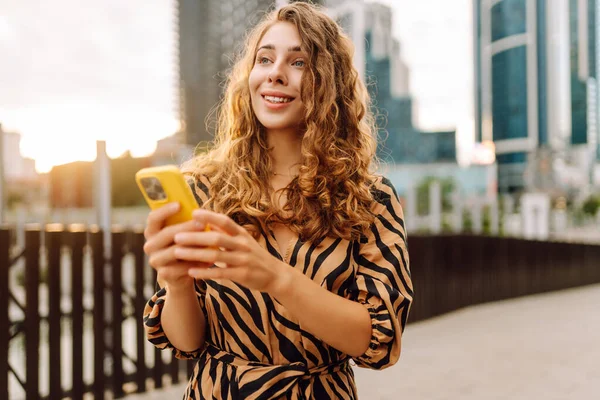 Elinde telefon olan genç bir kadın şehir manzarasının arka planında bir sokak arasında geziniyor. Güzel bir kadın telefonuyla güneşli bir günün tadını çıkarır. Teknoloji konsepti. Yaşam biçimi.