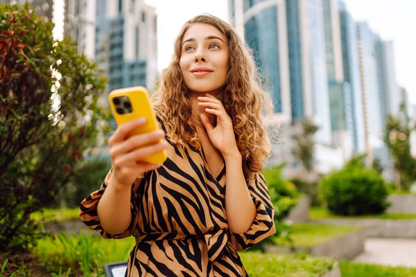 Elinde telefon olan genç bir kadın şehir manzarasının arka planında bir sokak arasında geziniyor. Güzel bir kadın telefonuyla güneşli bir günün tadını çıkarır. Teknoloji konsepti. Yaşam biçimi.