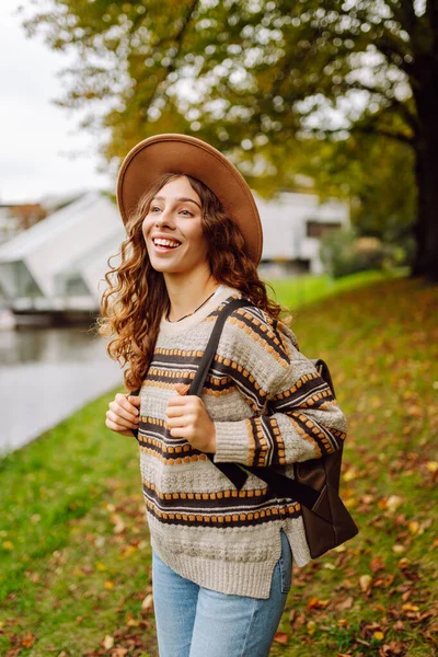 Sonbahar parkında yağmurluk ve şapkalı neşeli bir kadın bir nehrin kenarında duruyor. Sırt çantalı genç bir turist doğa ve doğa yürüyüşünün tadını çıkarıyor. Mevsimlik ve turizm kavramı.