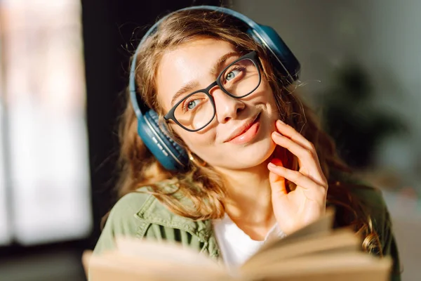 Kablosuz kulaklık takan ve güneş ışığında kitap tutan genç bir kadının yakın çekimi. Gözlüklü, gülümseyen bir kadın kitap okuyor ve rahat bir odada müzik dinlemekten hoşlanıyor. Konsept: eğitim, teknoloji.