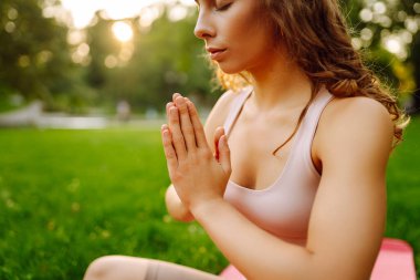 Genç bir kadın güneşli bir parkta yoga minderinde pratik yapıyor. Atletik bir kadın gün batımında dışarıda egzersiz yapar. Rahatlama, dinlenme ve aktif bir yaşam tarzı kavramı.
