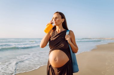Kumsalda elinde bir şişe suyla duran mutlu hamile kadın. Omzunda yoga minderiyle açık deniz manzarasının tadını çıkaran bir çocuk bekleyen sportif kadın. Annelik kavramı.
