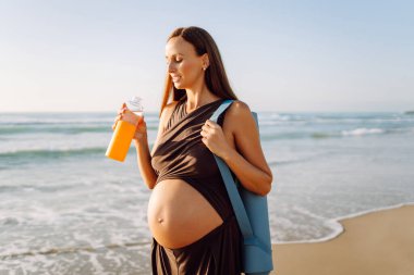 Kumsalda elinde bir şişe suyla duran mutlu hamile kadın. Omzunda yoga minderiyle açık deniz manzarasının tadını çıkaran bir çocuk bekleyen sportif kadın. Annelik kavramı.