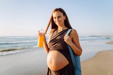 Kumsalda elinde bir şişe suyla duran mutlu hamile kadın. Omzunda yoga minderiyle açık deniz manzarasının tadını çıkaran bir çocuk bekleyen sportif kadın. Annelik kavramı.