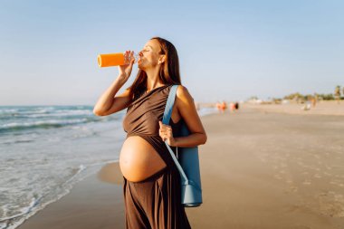 Kumsalda elinde bir şişe suyla duran mutlu hamile kadın. Omzunda yoga minderiyle açık deniz manzarasının tadını çıkaran bir çocuk bekleyen sportif kadın. Annelik kavramı.