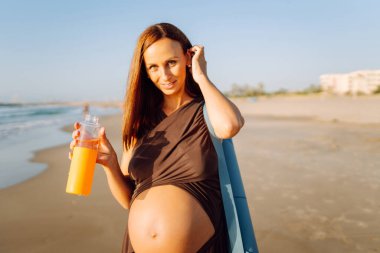 Kumsalda elinde bir şişe suyla duran mutlu hamile kadın. Omzunda yoga minderiyle açık deniz manzarasının tadını çıkaran bir çocuk bekleyen sportif kadın. Annelik kavramı.