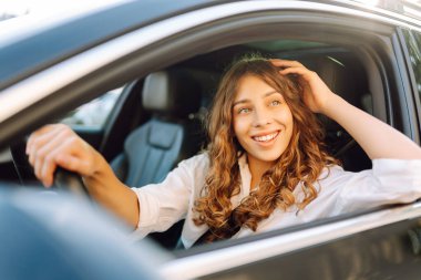 Arabanın direksiyonunda oturmuş pencereden dışarı bakan gülümseyen kadının portresi. Modern arabadaki kadın sürücü güneşli günün tadını çıkarıyor. Nakliye, taksi konsepti. Yaşam biçimi.