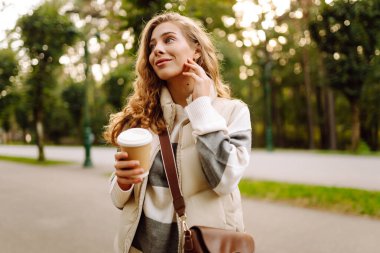 Parkta güneşli bir parkta elinde bir bardak sıcak içecekle bekleyen neşeli bir kadın. Genç bir kadın sonbahar yürüyüşünün tadını çıkarıyor ve sıcak bir şeyler içiyor. Boş zaman, hafta sonu konsepti..