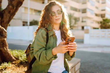 Güneş gözlüklü mutlu bir kadının portresi ve gün batımında parkta duran bir telefon. Güzel bir kadın eğleniyor, dışarıda telefonuyla mesajlaşıyor. Yürüyüş, eğlence konsepti. Yaşam biçimi.