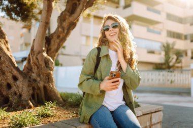 Güneş gözlüklü mutlu bir kadının portresi ve gün batımında parkta duran bir telefon. Güzel bir kadın eğleniyor, dışarıda telefonuyla mesajlaşıyor. Yürüyüş, eğlence konsepti. Yaşam biçimi.