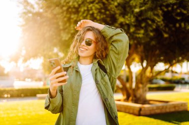 Güneş gözlüklü mutlu bir kadının portresi ve gün batımında parkta duran bir telefon. Güzel bir kadın eğleniyor, dışarıda telefonuyla mesajlaşıyor. Yürüyüş, eğlence konsepti. Yaşam biçimi.