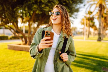 Güneş gözlüklü mutlu bir kadının portresi ve gün batımında parkta duran bir telefon. Güzel bir kadın eğleniyor, dışarıda telefonuyla mesajlaşıyor. Yürüyüş, eğlence konsepti. Yaşam biçimi.