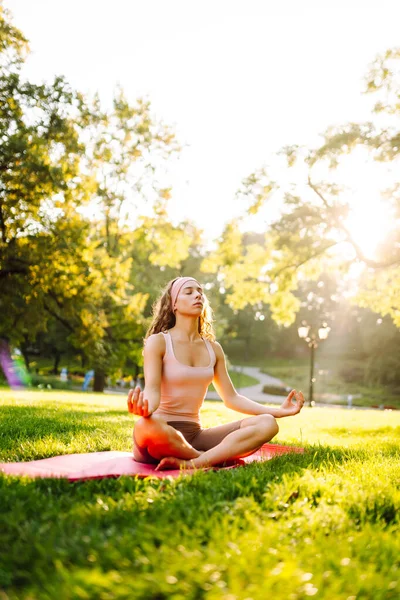 Genç bir kadın güneşli bir parkta yoga minderinde pratik yapıyor. Atletik bir kadın gün batımında dışarıda egzersiz yapar. Rahatlama, dinlenme ve aktif bir yaşam tarzı kavramı.
