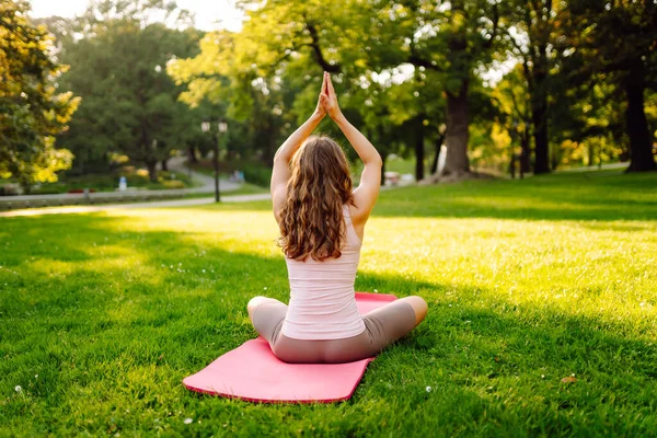 Genç bir kadın güneşli bir parkta yoga minderinde pratik yapıyor. Atletik bir kadın gün batımında dışarıda egzersiz yapar. Rahatlama, dinlenme ve aktif bir yaşam tarzı kavramı.