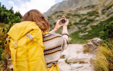 Sarı sırt çantalı kadın yürüyüşçünün dağ manzarasına pusula tutuşu. Pusula ile yön arayan ve vahşi doğayı keşfeden genç bir kadın. Doğa konsepti, navigasyon.