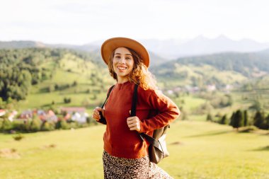 Dağlarla çevrili yemyeşil bir tarlada duran gülümseyen kadın. Sırt çantalı genç bir bayan gezgin dağ manzarasının tadını çıkarıyor ve temiz havada yürüyor. Rahatlama kavramı.