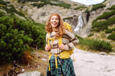 Güneşli bir günde dağ yolunda sırt çantalı güzel bir bayan yürüyüşçü. Şelale manzarasının tadını çıkaran güzel bir kadın, doğayı keşfediyor. Yürüyüş konsepti, vahşi doğa.