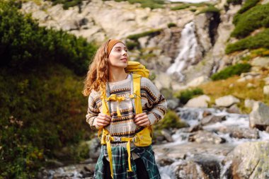 Güneşli bir günde dağ yolunda sırt çantalı güzel bir bayan yürüyüşçü. Şelale manzarasının tadını çıkaran güzel bir kadın, doğayı keşfediyor. Yürüyüş konsepti, vahşi doğa.