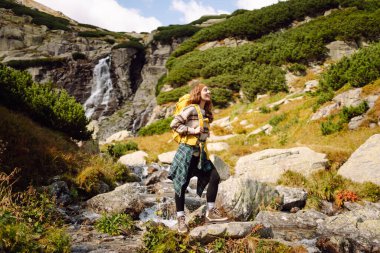 Güneşli bir günde dağ yolunda sırt çantalı güzel bir bayan yürüyüşçü. Şelale manzarasının tadını çıkaran güzel bir kadın, doğayı keşfediyor. Yürüyüş konsepti, vahşi doğa.