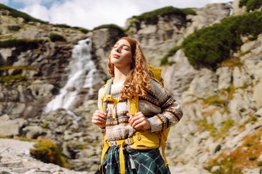 Güneşli bir günde dağ yolunda sırt çantalı güzel bir bayan yürüyüşçü. Şelale manzarasının tadını çıkaran güzel bir kadın, doğayı keşfediyor. Yürüyüş konsepti, vahşi doğa.