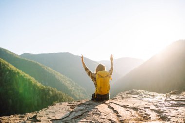 Parlak sarı sırt çantalı bir kadın yürüyüşçü bir uçurumda oturur ve dağ sırasına bakar. Genç kadın özgürlüğü hissediyor ve gün batımında dağ manzarasının tadını çıkarıyor. Özgürlük ve macera kavramı.
