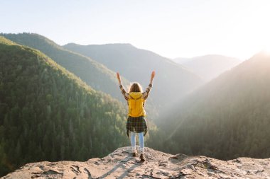 Parlak sarı sırt çantalı bir kadın yürüyüşçü bir uçurumda oturur ve dağ sırasına bakar. Genç kadın özgürlüğü hissediyor ve gün batımında dağ manzarasının tadını çıkarıyor. Özgürlük ve macera kavramı.