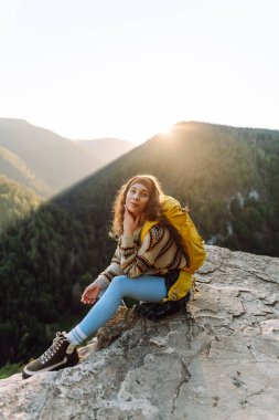 Parlak sarı sırt çantalı bir kadın yürüyüşçü bir uçurumda oturur ve dağ sırasına bakar. Genç kadın özgürlüğü hissediyor ve gün batımında dağ manzarasının tadını çıkarıyor. Özgürlük ve macera kavramı.