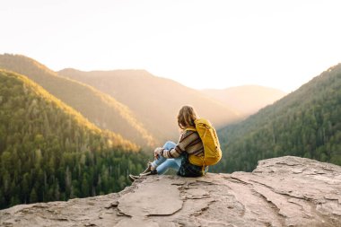 Parlak sarı sırt çantalı bir kadın yürüyüşçü bir uçurumda oturur ve dağ sırasına bakar. Genç kadın özgürlüğü hissediyor ve gün batımında dağ manzarasının tadını çıkarıyor. Özgürlük ve macera kavramı.