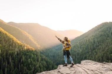 Parlak sarı sırt çantalı bir kadın yürüyüşçü bir uçurumda oturur ve dağ sırasına bakar. Genç kadın özgürlüğü hissediyor ve gün batımında dağ manzarasının tadını çıkarıyor. Özgürlük ve macera kavramı.