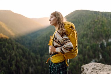 Parlak sarı sırt çantalı bir kadın yürüyüşçü bir uçurumda oturur ve dağ sırasına bakar. Genç kadın özgürlüğü hissediyor ve gün batımında dağ manzarasının tadını çıkarıyor. Özgürlük ve macera kavramı.