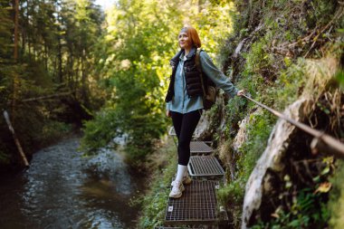 Sırt çantalı güzel bir bayan yürüyüşçü derenin yanındaki metal yürüyüş yolunda duruyor. Genç bir kadın güneşli ormanda vahşi doğanın tadını çıkarıyor. Doğa konsepti, yürüyüş. Etkin yaşam biçimi.